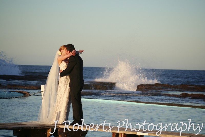 Bridal couple kissing at North Narrabeen Beach with breaking wave behind them - wedding photography sydney
